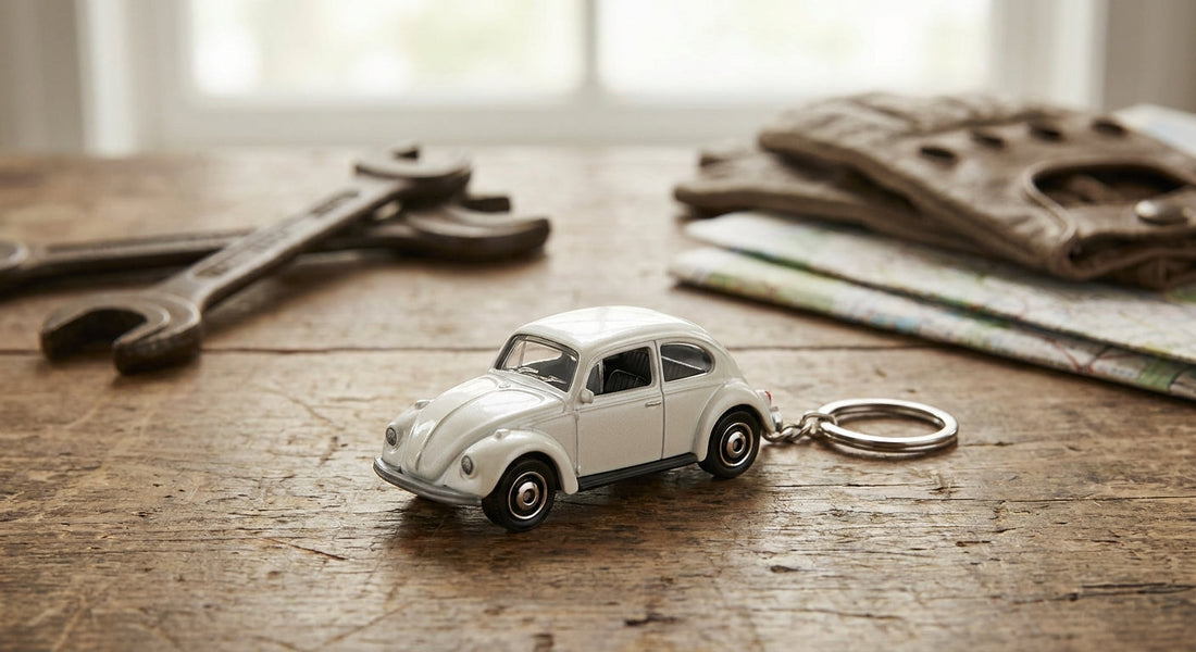 Detailaufnahme eines perlweißen Schlüsselanhänger VW Käfer als Modellauto auf einem Holztisch, umgeben von Oldtimer-Elementen.